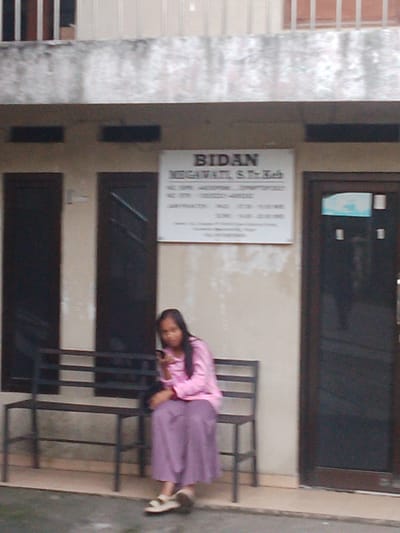 A woman in a pink top and purple skirt sits on a metal bench in front of a building. She is holding a mobile phone. A sign on the building reads "BIDAN MEGAWATI S.Tr.Keb". The building appears to be a clinic or clinic-like establishment in Megamendung, Indonesia, evidenced by the sign and the general architectural style of buildings in that region. The woman is looking down at her phone and is not interacting with anyone else in the scene. The surroundings are neutral, with a concrete walkway in front of the building.