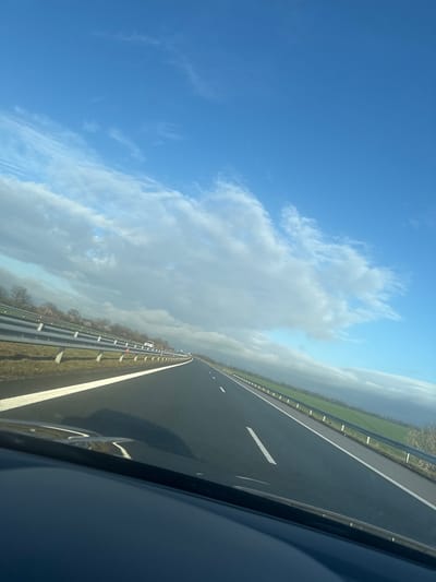 The image depicts a multi-lane asphalt highway on a clear day with scattered clouds, viewed from inside a moving vehicle. The location is specified as Vodenichane, Bulgaria.
The foreground shows the dark dashboard or hood of a vehicle occupying the bottom approximately 15% of the frame. The highway extends from the lower center towards the horizon, featuring dark asphalt. On the left side of the road, a solid white line marks the shoulder, followed by a wide solid white line, then a dashed white line indicating a lane divider. On the right side, a solid white line delineates the shoulder.
Metal crash barriers (guardrails) run parallel to the road on both sides. The barrier on the left consists of three horizontal rails supported by vertical posts, separating the highway from a strip of grass and then a line of leafless deciduous trees. The barrier on the right also features multiple horizontal rails, bordering a green field which extends to the distant horizon where a faint line of trees is visible. Several small orange traffic cones or markers are interspersed along the right crash barrier. In the distance on the opposing lanes (to the left of the central barrier), a white car is visible.
The sky occupies the upper portion of the image. The upper-right quadrant is clear blue. A band of white and light grey cumulus clouds stretches diagonally from the mid-left towards the far right horizon, with some darker grey cloud formations present on the right. No individuals are visible, though the perspective indicates a person is operating or riding in the vehicle from which the photograph was taken.