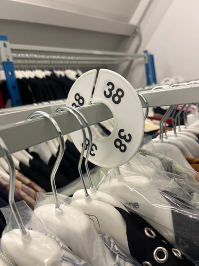 A close-up shot shows a clothing rack filled with garments on hangers. The garments are mostly obscured by plastic coverings. A white, circular size divider labeled "38" in bold black font is attached to the rack. The divider is split, allowing it to be easily added or removed. The rack itself is a light gray metal. The background includes other racks and clothing, suggesting a retail or storage environment.