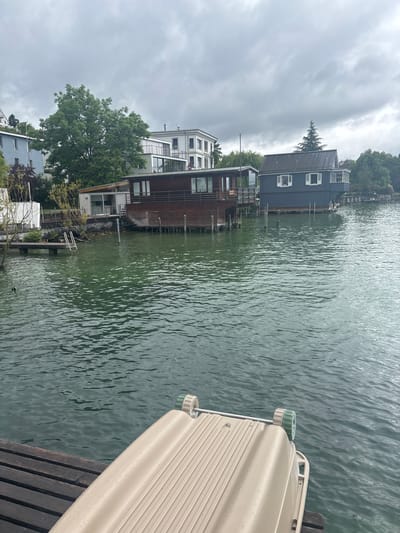 This image shows a waterfront scene in Kilchberg, Switzerland, likely taken from a dock or pier. The foreground contains part of a light-colored boat or floatation device resting on weathered wooden planks. The water is visible, showing a rippled surface. The backdrop features houses built on the water's edge and on land. There is a multi-story house with wood exterior, a blue-grey house with white framed windows. Above, there is a cloudy sky with overcast conditions.