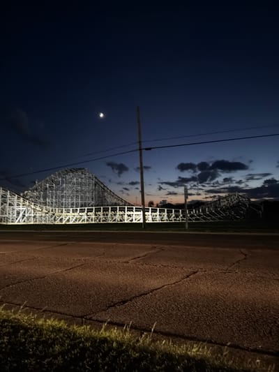 This image captures a twilight scene of a wooden roller coaster in Wisconsin Dells, United States. The sky is a deep, dark blue, transitioning to lighter shades of orange and pink near the horizon, indicating either sunrise or sunset. Wispy clouds are scattered across the sky, adding depth to the scene.

The roller coaster structure, primarily white, dominates the middle ground. Its intricate wooden framework is illuminated by artificial lighting, creating a stark contrast against the darkening sky. The tracks of the roller coaster are visible, winding and climbing, hinting at the thrilling ride it offers.

To the right of the roller coaster, a paved road is visible. The road appears to be cracked and somewhat worn, with dry grass and weeds growing along its edge. The lighting suggests that it is nighttime or very late twilight, as artificial lights are casting shadows and illuminating the road and the surrounding grass.

In the upper left quadrant of the image, a bright, crescent moon is visible against the dark sky, a prominent celestial object in the low-light environment. Power lines stretch across the sky, a common man-made element that often accompanies outdoor scenes.

The overall mood of the image is serene yet hints at excitement due to the presence of the roller coaster. The low light and the presence of the moon suggest a calm evening or early morning at an amusement park. No people are visible in the image, making the roller coaster and the natural elements of the sky and landscape the main subjects. There is no visible text.