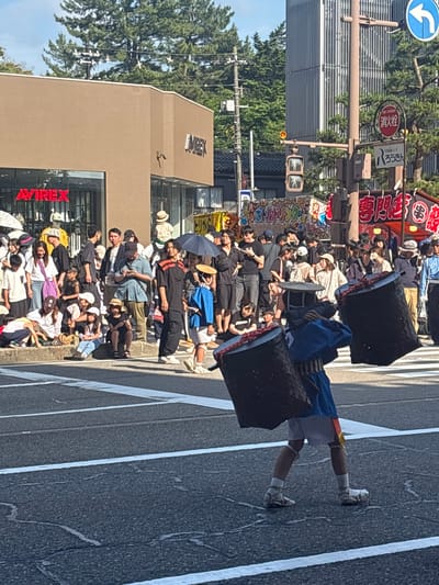 This is a daytime photograph depicting a street scene in Katamachi, Kanazawa, Japan, during what appears to be a public event or festival.
In the immediate foreground, a person in traditional Japanese attire is crossing the street from left to right on a white-striped crosswalk. They are wearing a blue short-sleeved garment with red-orange accents, white shorts, white tabi-style socks, and straw sandals. A broad, flat straw hat covers their head. They are carrying two large, dark, cylindrical, woven objects, resembling baskets or drums, suspended from a harness worn over their shoulders. These objects are positioned one on each side of their body.
In the mid-ground, a substantial crowd of people, casually dressed, lines the left side of the street. Many are standing, observing the event, while others are seated on the curb. Behind them, a two-story commercial building with a light brown facade is visible. The ground floor features large display windows and a red illuminated sign that reads "AVIREX." A separate, partially visible sign on the upper facade reads "IMREX."
Further to the right, a multi-post traffic light structure stands. It displays a circular blue sign with a white left-turn arrow, a circular red sign with white Japanese characters reading "消火栓" (Shōkasen - Fire Hydrant), and a white rectangular sign with Japanese text "はろうきん" (Haroukin - referring to Hokuriku Bank). Colorful banners with Japanese text, including "ボトルドリンク" (Bottle Drink) and "専門店" (Senmonten - specialty store), are strung across the street in the background, further indicating a festival atmosphere.
In the distant background, above the buildings and trees, a tall, modern building with a vertically ribbed, light-colored facade is visible under a clear blue sky. Green trees are interspersed throughout the scene, particularly in the upper right. The asphalt road shows white road markings and minor surface cracks. The scene is brightly lit by direct sunlight, casting distinct shadows.
