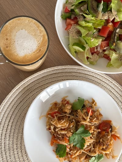 An overhead shot shows a meal laid out on a wooden table. In the foreground, a white plate holds a serving of rice pilaf garnished with fresh parsley. To the left, a glass mug contains a frothy coffee drink, possibly a cappuccino or latte. Behind the coffee and to the right, a white bowl is filled with a fresh green salad containing lettuce, tomatoes, and cucumbers, all dressed in a creamy salad dressing. The table itself has a visible wood grain. The image appears to be taken indoors, likely in a home or restaurant setting. There is no indication of the city of Sakmary, Russia.