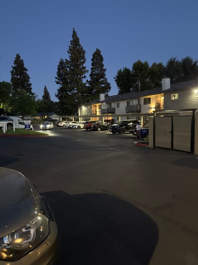 A twilight scene depicts an apartment complex parking lot with several vehicles. Tall coniferous trees are visible behind the buildings, and the sky is a deep blue, indicating evening. In the foreground, the hood and headlight of a silver car are prominent. The parking lot is paved, with a red-curbed island in the middle. Several cars are parked in rows, including a white van, a silver sedan, and a black SUV. Two individuals are walking near the apartment buildings. The buildings are two stories high, with balconies and exterior staircases. A large gate with horizontal slats is positioned on the right side of the frame. The overall impression is a residential setting at dusk.