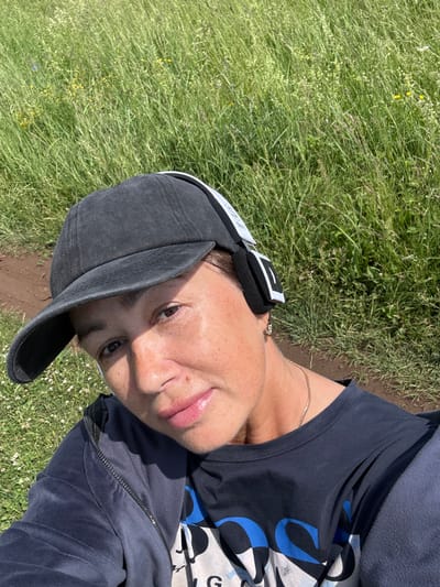 A medium shot selfie shows a woman wearing a dark baseball cap and headphones. She has on a dark blue t-shirt and a dark blue zip-up jacket. The woman is looking down and to her right with a slight smile. Her hair is dark brown and pulled back. The background consists of tall green grass and some brown dirt. The location is the city of Kalinin, Russia.