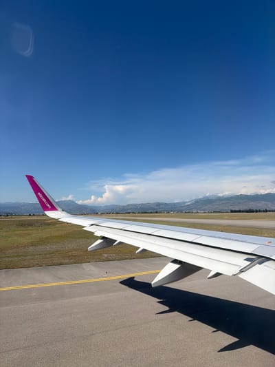 The image shows an airplane wing with the Wizz Air logo on the purple wingtip. The wing is positioned over an airport tarmac with yellow lines marking a runway. In the background, there are green fields, trees, buildings, and mountains under a clear blue sky with scattered clouds. The scene appears to be at the Podgorica Airport in Donja Cijevna, Montenegro. There are no people visible in the image. The airplane is either on the ground or taxiing, as indicated by the shadow of the wing on the tarmac.