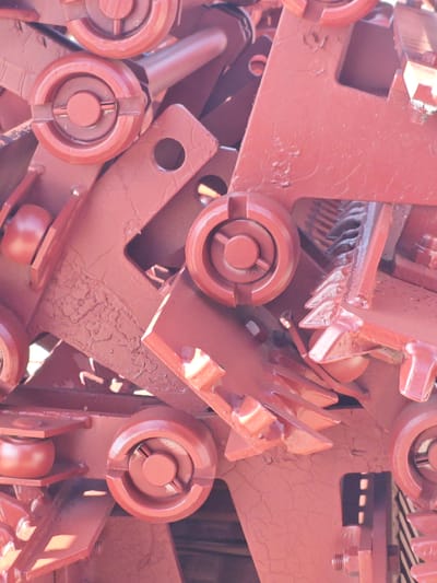 The image is a close-up, high-angle shot of a pile of weathered, red-painted metal machinery parts. The objects are densely packed, obscuring any clear background or distinct environment. The machinery appears to be industrial, possibly part of a large piece of equipment like a combine harvester or a construction machine, characterized by various circular components with notches, sharp-toothed elements, and angular plates. The paint is peeling and cracked in several areas, revealing the underlying metal and suggesting age and outdoor exposure. There are no people visible in the image, and no specific location details are discernible from the objects themselves. The scene does not depict any actions or interactions.
