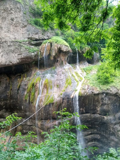 The image depicts a natural scene of a waterfall cascading down a rocky cliff face in Khushto-Syrt, Russia. The cliff is layered and appears to be composed of sedimentary rock, with patches of vibrant green moss and vegetation growing on its surface. Several streams of water flow down the cliff, some forming thin, ribbon-like waterfalls, while others appear as trickles. The surrounding environment is lush and green, with dense foliage and trees framing the top and foreground of the image. Sunlight filters through the leaves, casting dappled light on the scene. A thin, light-colored rope or cable is visible stretching horizontally across a portion of the waterfall. There are no people or animals visible in the image. The overall impression is one of natural beauty and tranquility.