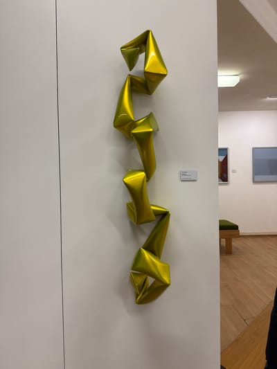 The image depicts a yellow abstract sculpture mounted on a white wall. The sculpture consists of angular, geometric shapes that are connected and arranged horizontally. Above the sculpture, there is a small, rectangular information sign. In the background, a portion of a building interior is visible, including a hallway or room with white walls, a doorway, and a section of wood flooring. Based on the information sign, the sculpture is a work by Wolfgang Stiller titled "Verwandlung" ("Transformation"), made of synthetic resin and car paint, and created in 2014. The location is likely an art gallery or museum. The city is Biberach an der Riß, Germany.