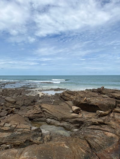 The image depicts a rocky shoreline at Ao Cho, Thailand. The foreground is dominated by large, textured brown rocks, some of which have small pools of water collected on their surfaces. Beyond the rocks, the turquoise ocean stretches to the horizon under a partly cloudy sky. Gentle waves crash against the shore and the scattered rocks. The scene is natural and serene, with no people or man-made structures visible, except for two small cairns made of stacked stones, one on the right side of the frame and another smaller one in the middle ground. The overall impression is one of a rugged and tranquil coastal landscape.