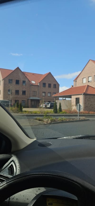 The image shows a view from inside a car, likely taken in Rietberg, Germany based on the buildings. Outside the car, there are several multi-story brick buildings with red roofs. A road with a sign runs alongside the buildings. The sky is blue with a few clouds. Inside the car, parts of the dashboard and steering wheel are visible, indicating a driver's perspective.