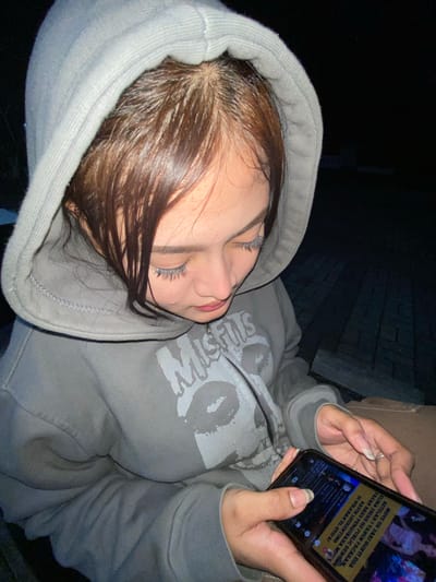 A medium shot captures a young woman wearing a grey hooded sweatshirt, seated in an outdoor setting at night. The woman is looking down at her phone, which she holds in her hands. Her hair is brown and appears wet. The background consists of dark, paved ground and hints of wooden structures. The scene is dimly lit, suggesting it is dark outside. The woman is the sole subject of the image. The presence of a wooden bench and paved ground suggests the location is likely a park or an outdoor public space within Mekar Bakti, Indonesia.