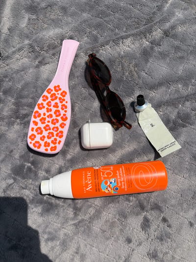 This media file contains an overhead shot of several personal care items laid out on a grey, textured blanket. The objects include a pair of tortoiseshell sunglasses, a small white rectangular object that appears to be an earbud case, a tube of hand cream labeled "SISTER'S AROMA SMART HAND CREAM," a pink Tangle Teezer hairbrush with an orange and pink leopard print pattern, and an orange spray bottle of "Avène EAU THERMALE SPF 50 Spray for children." The spray bottle has text in both French and English indicating "VERY HIGH PROTECTION" and "VERY WATER RESISTANT" for sensitive children's skin. The lighting suggests it is daytime, possibly outdoors, with a shadow visible in the lower right corner. The arrangement of the items implies they are ready for use or have just been used, possibly in preparation for or after a day in the sun. The scene is static, with no people or active event depicted.