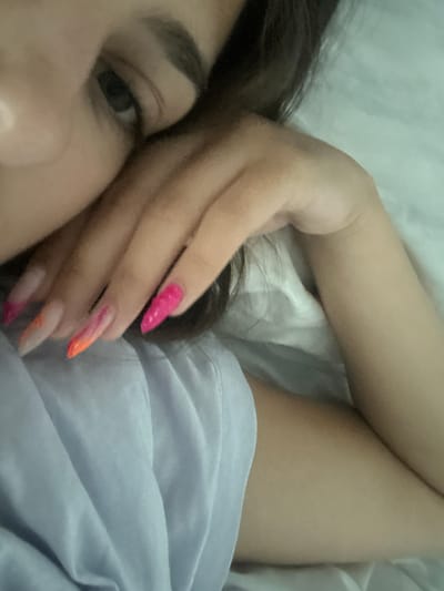 The image shows a close-up of a person's face and hand. The person is lying on a white surface, possibly a bed or sofa, and appears to be resting their head on their hand. Their eyes are partially visible, and they have dark hair. The hand has long, acrylic nails painted in a gradient of pink and orange. The background consists of white bedding and a blue garment. The image is taken indoors, and the lighting is soft and diffused. There is no visible information to place this image in Stațiunea Zoologică Marină Agigea, Romania.