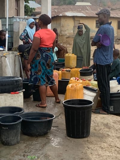 A group of people are lined up near water taps. Some are filling containers, while others wait their turn. Buckets and yellow jugs are stacked in rows, waiting to be filled. One man holds a purple scoop. A woman in an orange shirt and blue patterned skirt stands at a tap. Another woman with a blue headscarf sits nearby. The setting appears to be a community water source.