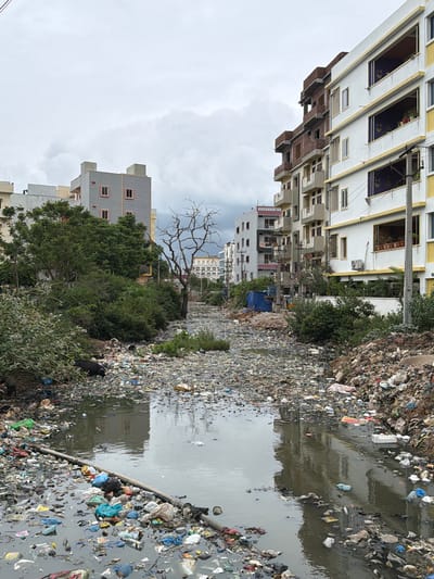 The image depicts a scene of severe environmental pollution in Shamshabad, India. A narrow, murky canal runs through the center of the frame, choked with a vast amount of garbage. Plastic bags, bottles, wrappers, and various other debris are submerged in the stagnant water and piled high on its banks. On the left side of the canal, lush green trees and bushes contrast sharply with the surrounding filth. Beyond the trees, modern multi-story residential buildings with balconies and windows are visible, indicating an urban or semi-urban setting. Some of these buildings appear to be under construction. Two dark-colored dogs are present near the canal's edge, foraging through the waste. The sky is overcast with heavy clouds. The overall impression is one of neglect and a severe lack of waste management in a developing urban area.