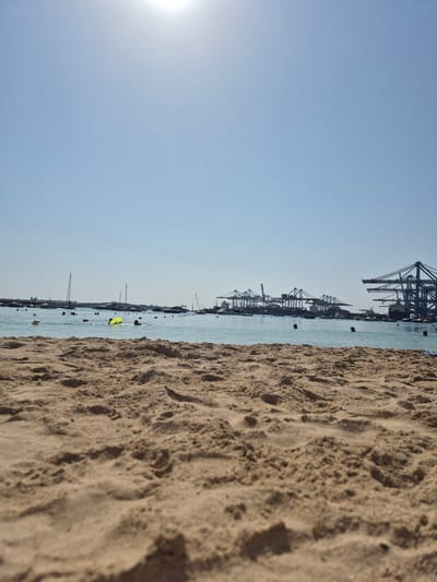 The image depicts a sunny day at a beach in Birżebbuġa, Malta, with the foreground showing detailed textures of sand. In the middle ground, the clear blue sea is populated by several people swimming and one person on a yellow float, along with various boats and buoys. The background reveals a distant industrial port with numerous large cranes, characteristic of the Marsaxlokk Freeport, a significant maritime facility in Malta. The sky is bright blue with a faint sun visible.