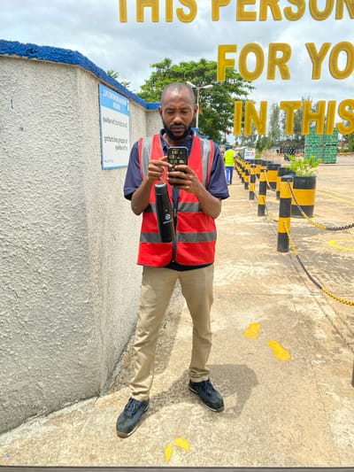 A Black male, appearing to be in his late 30s or early 40s, stands on a concrete walkway. He is wearing a navy blue short-sleeved shirt, a high-visibility red vest with reflective stripes, and tan-colored trousers. On his feet are dark blue athletic shoes. He holds a black smartphone in his right hand and a black insulated water bottle with a logo in his left hand. The man is looking down at his phone.
Behind him, to the left, is a textured white concrete wall topped with blue coping. A sign is attached to the wall, featuring text in an upside-down, reversed orientation. Further to the right, a chain-link fence with alternating black and yellow bollards and chains separates the walkway from an area with stacked green crates. In the background, trees with green foliage and a cloudy sky are visible. The overall setting appears to be an industrial or logistical facility. The sign on the wall suggests the location is in Kinkinau, Nigeria.
