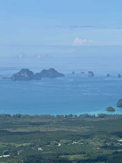 The image shows a panoramic view of the coastline near Ban Din Daeng Nui, Thailand. In the foreground, a dense forest covers the land, with some buildings scattered within. The forest transitions to a strip of trees along the coast, which meets a vast expanse of blue ocean. The ocean is dotted with several karst islands, some of which are covered in vegetation. The sky is a pale blue with a few scattered clouds. The overall scene is tranquil and natural, showcasing the tropical landscape of the region.