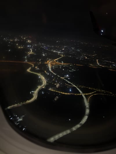 An aerial view through an airplane window at night showcases a city, Yufengshan, China, with winding roads or highways illuminated by streetlights. City lights of varying intensities dot the landscape, indicating urban areas. In the upper-left corner, a portion of the airplane's wing is visible. The scene is characterized by a low-light, nighttime environment.