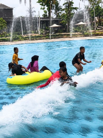 In the city of Eampat Kwalamencirim, Indonesia, a group of children are enjoying a water park. The scene is set in a clear blue pool with several fountains spraying water in the background. In the foreground, a yellow inflatable tube is floating in the water, with a girl in a purple hijab and a girl in a black swimsuit seated on it. Another girl in a black swimsuit is partially submerged in the water, next to the tube. To the right, a boy in a black t-shirt and shorts is standing in the water, looking to his left. Further to the right, a girl in a red swimsuit is partially visible, her face obscured by a wave. A boy in a yellow shirt is visible in the upper left portion of the frame. The overall atmosphere is one of playful activity and enjoyment in a water recreation setting.
