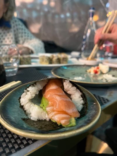 The image captures a close-up, top-down view of a meal, likely in a restaurant setting. The primary focus is a single sushi hand roll, or temaki, presented in an artisanal blue-green ceramic bowl on the right side of the frame. This hand roll consists of white sushi rice and green avocado slices, generously topped with fresh, light orange salmon. The ingredients are enclosed within a dark sheet of nori (seaweed).

In the background, to the left, another plate is visible with several out-of-focus sushi or maki rolls, along with what appears to be a small portion of wasabi and pickled ginger. A person's hands are visible in the upper left, holding chopsticks, suggesting they are in the process of eating or about to pick up food. Reflections from overhead lights are scattered across the table surface, indicating a dimly lit indoor environment, possibly an evening meal. A patterned placemat is partially visible beneath the foreground plate. The scene conveys a relaxed dining experience. There is no visible text in the image.