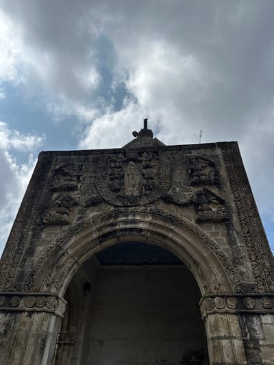 This is a low-angle, exterior, full-shot photograph of the facade of the San Andrés Apóstol Catholic Church in San Andrés Calpan, Mexico. The church's facade is made of stone and features intricate carvings, including a large central crest, winged figures, and decorative borders. An arched entryway is prominent in the lower portion of the image. The sky above is partly cloudy with a mix of white and gray clouds. The lighting suggests it is daytime. The image is focused on the architectural details of the church.