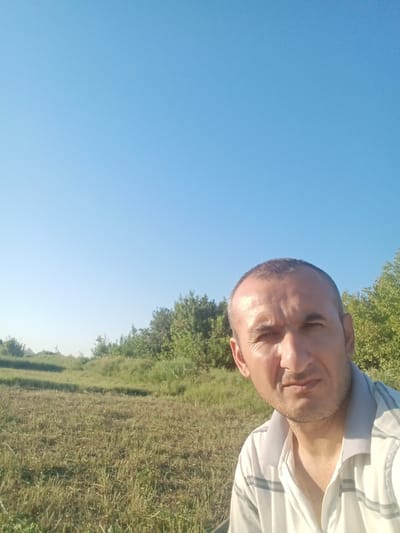 The image is a selfie taken outdoors with a clear blue sky dominating the upper portion. In the foreground, a man with short, dark hair and a light beard is visible from the chest up. He is wearing a white polo shirt with thin grey horizontal stripes. His expression is neutral. The background depicts a rural landscape with dry, cut grass in the immediate vicinity. Further back, there are rolling hills covered in green vegetation and trees, with some structures that appear to be houses or farm buildings in the distance. The lighting suggests it is daytime, likely late morning or early afternoon, given the brightness and lack of strong shadows. The overall scene conveys a sense of open space and a rural setting. Based on the provided information, this scene is located in the city of Romitan, Uzbekistan.
