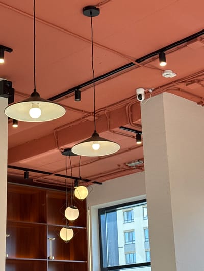 The image shows a collection of ceiling lights and security cameras mounted on a pink wall. The lights vary in design, with some being track lighting with small, cylindrical fixtures, while others are pendant lamps with wide, conical shades. One set of pendant lamps features multiple spherical bulbs attached to a wire frame. A white security camera with a lens and a small white base is visible on the upper left side of the wall. Another small, white circular device, possibly a smoke detector or another sensor, is mounted above it. The wall is covered in electrical conduits and wiring, suggesting an interior space with exposed infrastructure.
In the background, a window reveals a glimpse of an urban environment with a building featuring multiple windows. The time of day appears to be daytime, as indicated by the natural light entering through the window. The overall impression is of a modern interior space, possibly an office, retail store, or public building, that incorporates industrial design elements. There is no visible text in the image.