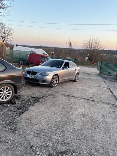 The scene depicts an outdoor, semi-urban environment in the city of Septemvritsi, Bulgaria, characterized by a deteriorated concrete and gravel surface. The time of day is either early morning or late afternoon, indicated by the sky's gradient from pale blue overhead to a light orange-pink near the horizon.

A silver BMW E60 5-Series sedan is centrally positioned, parked facing forward on the rough ground. Its front-end is partially obscured on the left by a dark-colored sedan, likely an older BMW (E36/E46 generation), of which only the rear driver's side wheel with a five-spoke silver alloy rim and a portion of the body panel are visible. The silver BMW displays a Bulgarian license plate, "B 5808 XB," signifying Sofia City registration, and is fitted with aftermarket multi-spoke alloy wheels.

The ground surface consists of heavily worn concrete mixed with loose gravel and dirt, showing significant cracking and exposed rebar in various sections. Multiple discarded cigarette butts are scattered across the ground in the foreground.

In the mid-ground, a green metal fence spans horizontally, delineating the immediate parking area from a background plot. Behind this fence, a red van and a white van are parked. To the far right, another segment of a green metal fence or gate is visible.

The background features several bare deciduous trees, suggesting a late autumn, winter, or early spring season. Distant low-rise buildings or a settlement are discernible on the horizon line, set against what appears to be a natural landscape of rolling hills. No individuals are present or engaged in any activity within the captured frame.