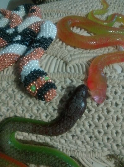 The image displays a collection of toy snakes arranged on a woven surface, likely a rug or placemat. There are at least three distinct toy snakes visible. One snake, in the foreground and to the left, appears to be a beaded craft, possibly a decorative item. It has a head with green bead eyes and is colored with white, black, and copper beads. To its right, a translucent orange and yellow snake coils across the woven background. In the lower right quadrant, another translucent snake, this one colored red and orange, has its head close to the beaded snake's body. A fourth snake, predominantly green with a dark brown patterned body, curves across the bottom of the image. The overall scene is a still life arrangement of these snake toys. There is no indication of people, text, time of day, weather, or specific location cues beyond the general setting of a surface with a woven texture.