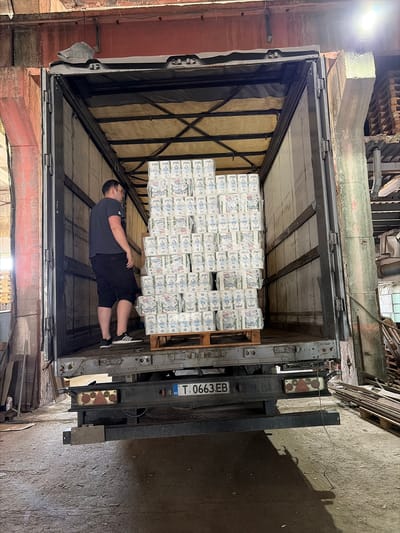 A photograph, oriented horizontally, depicts the rear of a truck filled with stacks of packaged toilet paper, with a man lying underneath a pallet of the product. The scene appears to be within a warehouse or loading dock area. The truck's license plate, which reads "T.0663.EB," indicates the country code BG, identifying it as Bulgarian.
The interior of the truck is visible, revealing multiple stacks of wrapped toilet paper on a wooden pallet. A man, wearing a dark t-shirt and black shorts, lies on the truck bed beneath the suspended pallet. He is facing toward the rear of the truck, with his feet near the liftgate.
The surrounding environment shows warehouse-like infrastructure, including exposed beams and weathered walls. The lighting is a mix of natural light from the truck's opening and artificial light sources within the warehouse. Given the information available, the location is presumed to be somewhere in Bulgaria. Further, given the presence of the Bulgarian license plate and that the truck appears to be being loaded/unloaded at an industrial facility, and based on available data, it is most likely located in the city of Dulovo, Bulgaria.