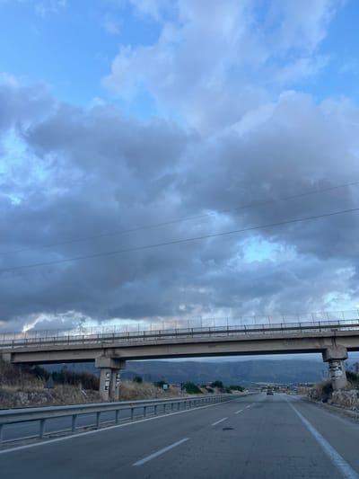 The image captures a view from a vehicle traveling on a highway in Manfredonia, Italy, under a cloudy sky. In the foreground, the highway stretches ahead with clear lane markings and a metal guardrail on the right. A bridge, supported by concrete pillars, spans across the highway. The bridge is equipped with a mesh fence along its sides. The landscape on either side of the highway is dry and sparse, with hills and mountains visible in the background. The sky is filled with a mix of dark and light clouds, suggesting an overcast or changing weather condition. There are a few cars visible on the highway in the distance.