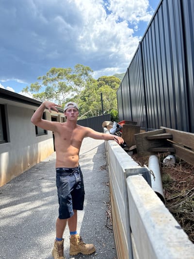 A shirtless young man, wearing a backwards baseball cap, navy shorts, and work boots, stands on a concrete path. He flashes a peace sign with his left hand and holds his phone in his raised right hand, presumably taking a selfie. Behind him, a grey fence, some trees, a house, and a cloudy sky are visible. The photo appears to be taken outside on a sunny day.