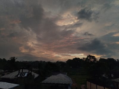 This image presents a serene, atmospheric landscape scene captured during either sunrise or sunset, likely over a semi-urban or rural area in Monrovia, Liberia.

The upper two-thirds of the frame are dominated by a vast, cloud-filled sky. Dense, multi-layered clouds in various shades of grey, dark blue, and muted purple stretch across the horizon. Hints of soft pink, orange, and pale yellow light are visible filtering through the clouds, particularly towards the center and right, indicating the low position of the sun. The heavy cloud cover contributes to a subdued and contemplative mood.

Below the sky, a landscape of dense tropical vegetation and scattered human settlements unfolds. A prominent band of dark green trees and bushes forms a thick silhouette across the mid-ground, creating a natural horizon line. In the foreground and mid-ground, several buildings are visible, characterized by their dark, corrugated metal roofs. One structure in the lower left features a lighter section of roofing, possibly reflective metal, while another in the lower right shows a textured wall surface.

No people are visible in the frame, and there is no discernible text. The overall impression is one of peaceful twilight, with the overcast conditions and the soft light creating a tranquil and slightly melancholic ambiance over the densely vegetated landscape.