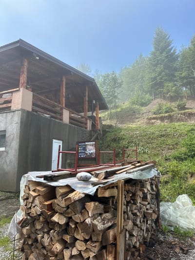 The image depicts an outdoor scene in Trešnjevik, Montenegro, featuring a rustic wooden structure with an extended porch or balcony, and a large pile of firewood in the foreground. The structure appears to be made of rough-hewn timber, with exposed wooden beams and a metal roof. A grey concrete wall forms the base of the building, with a white door and a window visible.

In front of the building, a substantial stack of chopped firewood is neatly arranged, covered with a translucent plastic sheet. Some wooden planks and a large grey rock rest on top of the firewood. A red metal frame, possibly part of a temporary structure or support, is positioned behind the firewood pile.

A sign with text and images is attached to the red metal frame. The surrounding environment is hilly and covered with dense green vegetation, including numerous trees, suggesting a rural or mountainous setting. The sky is a light blue with some atmospheric haze or fog, particularly visible between the trees on the hillside. The overall impression is one of a remote, possibly rural dwelling or facility in a natural landscape. There are no people visible in the image.