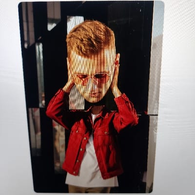 A portrait-oriented image depicts a single individual, likely a child or young adolescent, positioned centrally against a dark, predominantly black background. The individual has short, reddish-brown hair and wears red-tinted, round-frame sunglasses. Both hands are raised to the sides of the head, pressing against the ears in a gesture. The person is dressed in an open, red, possibly corduroy or ribbed, button-up jacket with two chest pockets, over a plain white t-shirt. Light-colored trousers or pants are faintly visible at the bottom of the frame. The background includes subtle reflections, suggesting dark, polished, or mirrored surfaces, with some geometric reflective elements visible on the left. Strong, uniform vertical light streaks are present across the entire image, notably over the subject's face and body, consistent with a photograph taken of a digital screen. There are no discernible visual cues within the image to identify its specific location within Gimpo-si (김포시), South Korea.