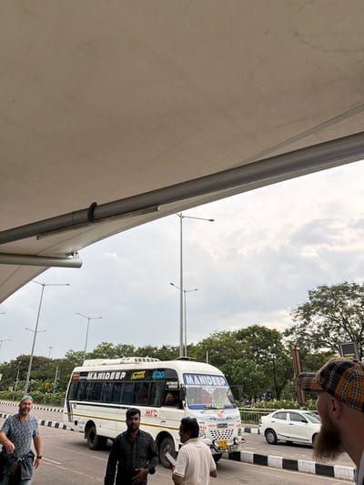In the city of Gollapalle Khurd, India, under a large, light-colored awning supported by metal beams, a white bus with "MANIDEEP Travels" written on its side is parked on a paved road. The bus has a blue stripe along its side and "AC COACH" printed on the front. Several people are visible in the scene. A man wearing a patterned t-shirt and a blue cap is walking towards the left, with a bag slung over his shoulder. Another man in a dark checkered shirt stands in the foreground, facing the camera. A third person, wearing a light-colored shirt, is partially visible to the right, holding a phone. The bus has its headlights on, and a person can be seen in the driver's seat. In the background, streetlights and trees line the road, and a white car is parked further down. The sky is overcast with clouds.