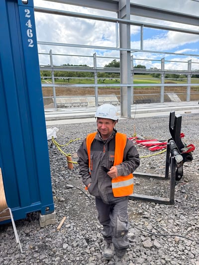 A man in a construction site is standing in front of a blue shipping container. He is wearing a white hard hat with writing on it, a grey work jacket, an orange reflective vest, and grey work pants. He is holding a small black object in his right hand. The ground is covered in gravel and there are construction materials scattered around. In the background, there is a building under construction with steel beams and a cloudy sky. The shipping container has the numbers "2462" written vertically on its side. The scene is outdoors, and the environment appears to be a construction site in Jodłownik, Poland.
