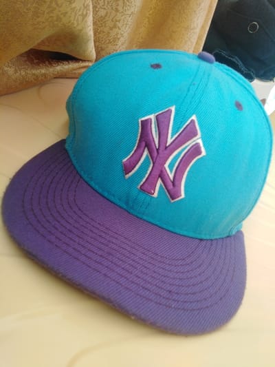 This image depicts a two-tone baseball cap positioned at an oblique angle on a light-colored flat surface, likely within an indoor environment located in Sigona ward, Kenya. The cap features a cyan-blue crown with a prominent purple "NY" logo, outlined in white, embroidered on its front. The brim is purple, exhibiting multiple parallel stitching lines. Visible details on the crown include purple ventilation eyelets and a central purple top button. The fabric of the cap, particularly the brim, shows minor superficial wear. The background consists of a golden-beige patterned fabric in the upper left and a partially visible dark blue or black fabric object with a metallic eyelet in the upper right. No individuals are present, and no actions or interactions are depicted.