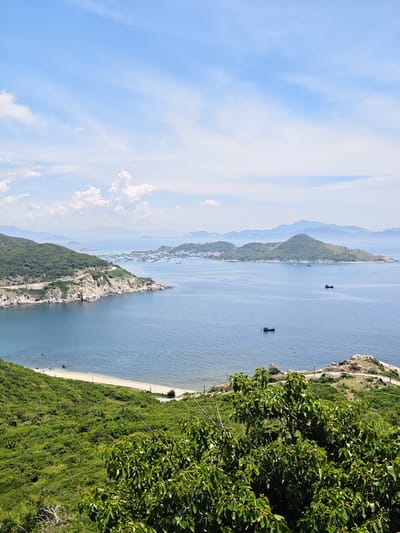 This image depicts a panoramic view of a coastal landscape, likely in Vinh Hai, Vietnam, featuring lush green hills, a tranquil blue sea, and a scattering of boats. The foreground is dominated by dense, verdant foliage, suggesting a hillside perspective. A winding road snakes along a rocky, tree-lined incline in the mid-ground. Beyond this, a sandy beach meets the calm, blue waters of the ocean. Further out, several islands dot the horizon, with a cluster of buildings and a harbor visible on one of them, hinting at a populated coastal town. A few boats are present on the water, one closer to the shore and others further out towards the islands. The sky is a bright blue, with scattered white clouds adding depth to the scene. The overall impression is one of natural beauty and serene coastal life.