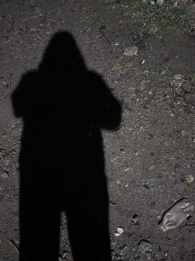 The scene depicts a prominent, dark, human-shaped shadow cast onto an uneven, unpaved ground surface in Lehvaz, Armenia. The shadow, consistent with an individual wearing a hooded garment, shows the subject's arms bent at the elbows, suggesting they are holding a device forward, likely a camera or mobile phone, to capture the image. The ground consists of compacted dirt, loose gravel, small stones, and various scattered debris, including what appears to be cigarette butts and dry organic matter. In the lower right portion of the frame, a crumpled, translucent plastic bottle lies horizontally on the ground. Sparse patches of green vegetation are visible along the upper right edge of the immediate environment. The lighting conditions are low, indicating an artificial light source positioned behind the subject casting the distinct shadow.