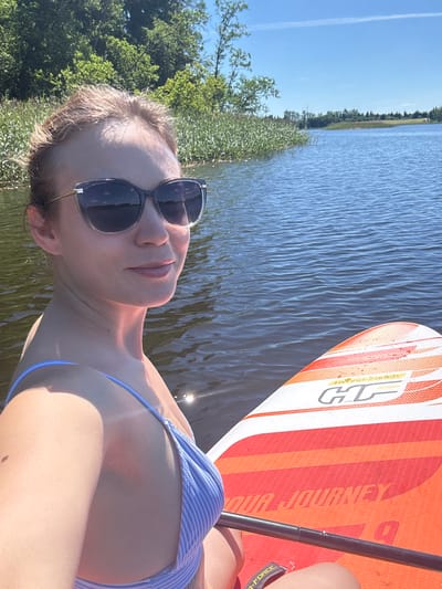 In the city of Lendži, Latvia, a woman with blonde hair pulled back and wearing sunglasses and a blue and white striped bikini, is paddleboarding on a body of water. She is positioned in the foreground, facing slightly to her right, with the water and distant trees in the background. The paddleboard, with orange and white detailing and the "HF JOURNEY" logo, is visible in the lower right portion of the frame. The sky is clear blue with a thin white cloud streak.