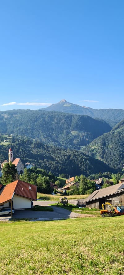 This is a vertical, eye-level, medium shot of a scenic mountain landscape in Deutschnofen - Nova Ponente, Italy. The foreground features a grassy hill sloping upwards, with some weeds and sparse dry grass. A paved driveway curves through the scene, leading to a house on the left with a red-tiled roof. To the right of the driveway, a rustic wooden barn stands, with a small yellow tractor and a colorful playground structure visible in front of it.

In the middle ground, a village is nestled amongst lush green trees. A church with a red-domed tower is prominent, with other buildings and houses scattered around it. The background is dominated by a majestic mountain range covered in dense evergreen forest, with a prominent, conical peak rising above the others in the distance. The sky is a clear, bright blue with a few wispy white clouds. The lighting suggests it is a sunny day, likely in the late morning or early afternoon. There are no people visible in the image.