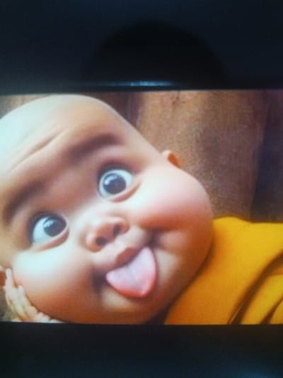 A close-up shot reveals a cartoonish, bald baby with a mischievous expression. The baby's eyes are wide and exaggerated, and its tongue is sticking out. The cheeks are chubby and flushed. The baby is wearing a yellow garment. An adult's shoulder is partially visible in the background. The lighting casts a shadow above the image, indicating it might be a photograph of a screen.