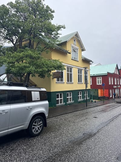 This image captures a street scene in Reykjavik, Iceland, on an overcast and wet day, specifically depicting a section of Austurstræti.
In the foreground, the rear left side of a silver, modern Land Rover Defender SUV is prominently visible. The vehicle is wet, indicating recent rain, and features a Thule roof rack. The asphalt street below is dark, wet, and speckled with white aggregate, with visible streaks of water and reflections on its surface.
The midground is dominated by two colorful multi-story wooden buildings. On the left, a two-story building is painted bright yellow with a dark green base and white trim around its numerous rectangular windows. It has a light green corrugated metal roof. A brown wooden sign with white lettering, reading "La Havana Club," is affixed to its facade. A large deciduous tree with dense green foliage partially obscures the upper left portion of this building.
To the right, behind the yellow building and slightly further down the street, stands a two-story red wooden building, also with white trim and a light green corrugated metal roof. This building also features multiple white-framed windows. A sign partially visible on its ground floor includes the words "Gin..." and "Kaldi." A red and blue circular "no entry" road sign is positioned on the sidewalk near this building. Two pedestrians are visible walking on the sidewalk in front of the red building, appearing to move away from the viewer.
Between the yellow and red buildings, a dark lamppost stands on the sidewalk. A green electric kick scooter is parked upright on the sidewalk near the red building. The sky is uniformly overcast and light grey, suggesting a fully cloudy day. The overall environment is one of a bustling city street after rain, characterized by distinctively colored Nordic architecture.