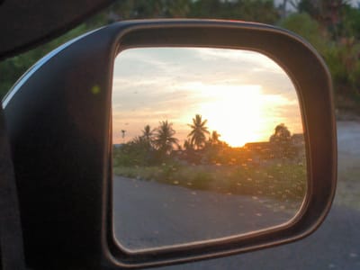 The image shows a side mirror of a vehicle reflecting a sunset scene in Cot Buket, Indonesia. The sun is setting behind a line of palm trees and vegetation along a road. The mirror also reflects a portion of the road and a hint of residential buildings obscured by the bright sunlight. The mirror itself is black and has water droplets on its surface. The vehicle the mirror is attached to is partially visible on the left.