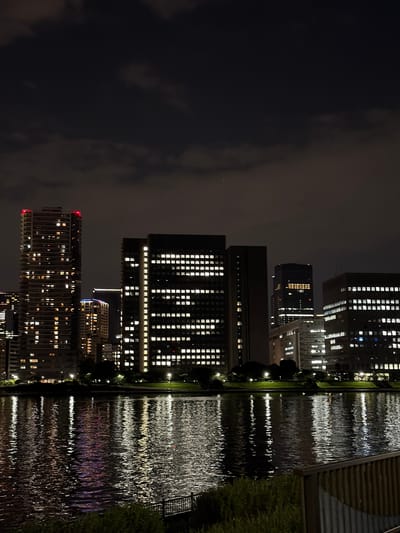 A nighttime cityscape in Koto, Japan, is captured from across a body of water. Tall, illuminated buildings dominate the background, their windows glowing with a warm, white light. A prominent skyscraper on the left features a red illuminated top. A building in the center has a pattern of illuminated squares forming a grid-like design. The water in the foreground reflects the lights of the city, creating shimmering streaks of color. The sky above is dark with scattered clouds. The foreground includes a dark, grassy embankment and a portion of a wooden railing. There are no visible people or animals in the scene.