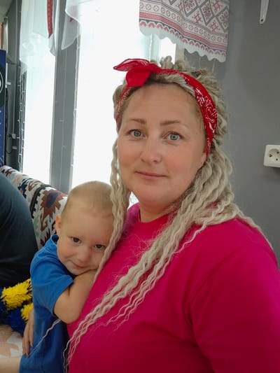 In the city of Cheremushki, Russia, a woman with blonde dreadlocks and a red bandana is holding a young child. The woman is wearing a pink shirt, and the child is wearing a blue shirt. They are sitting on a couch with a patterned fabric. In the background, there are curtains with embroidered designs and a wall with a white electrical outlet. The lighting suggests it is daytime, with light coming from a window. The scene appears to be indoors, likely in a residential setting.
