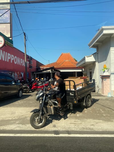 The image depicts a street scene in Ngrame, Indonesia, under a clear blue sky. In the foreground, a man wearing a black t-shirt, black shorts, and a black baseball cap is riding a black three-wheeled vehicle. The vehicle has a platform in the back loaded with cardboard boxes. The man is looking forward, and his left hand is on the handlebars. To the left of the vehicle, a dark-colored car is parked. Behind the car and the three-wheeled vehicle, several motorcycles are parked. A large red building with white lettering that reads "NIPPON PAINT" is visible on the left side of the image. The building has several air conditioning units mounted on its exterior. Further back, traditional Indonesian roofs with orange tiles are visible. To the right, a white building with a decorative pillar displaying the number "67" and an ornamental yellow object resembling corn on the cob is present. The ground is paved with concrete, and white lines indicating a crosswalk are visible in the lower portion of the image. Overhead power lines are also a prominent feature of the scene.